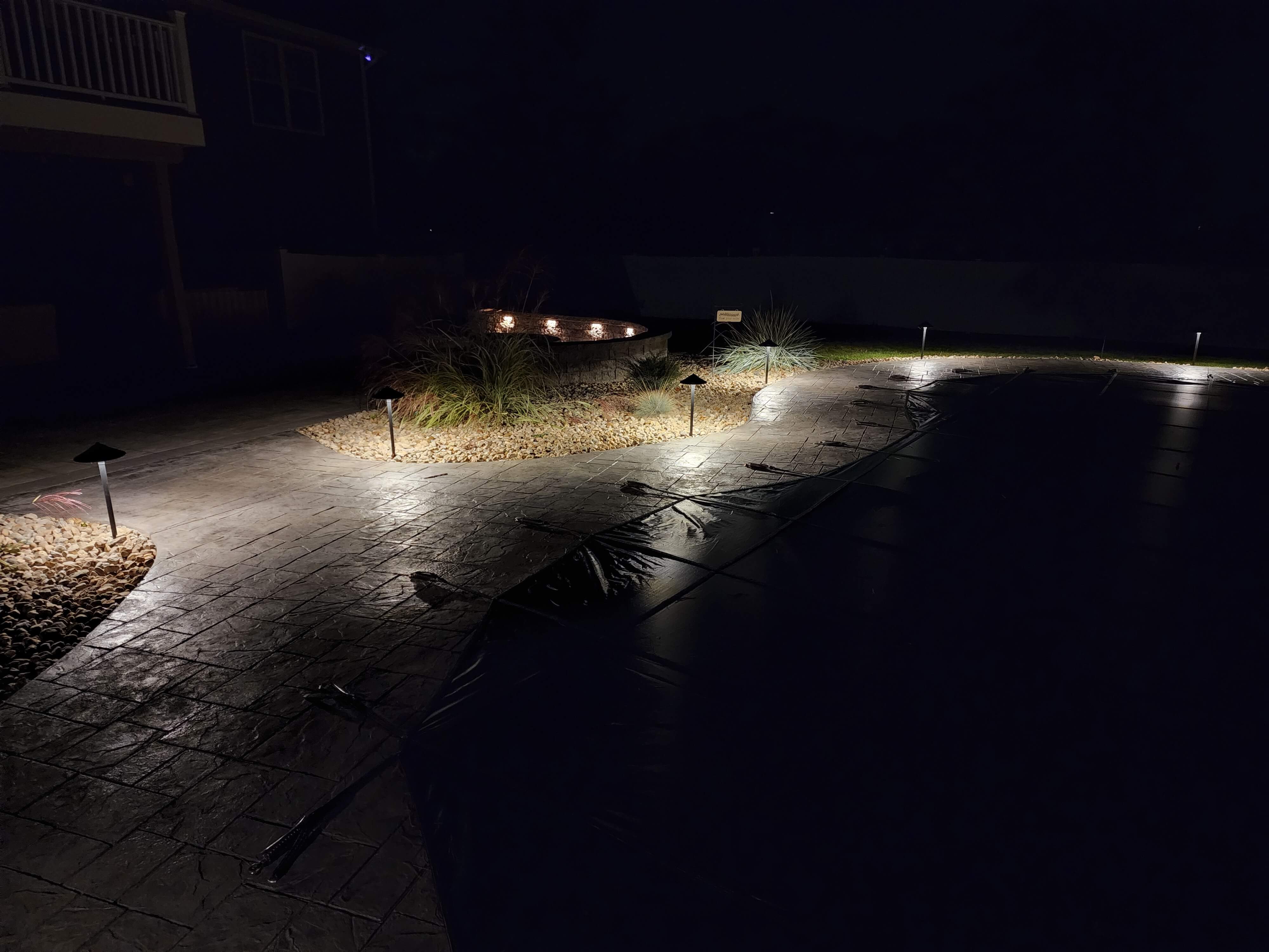 Pool patio at night showing pathway lights with fire pit wall lighting in background